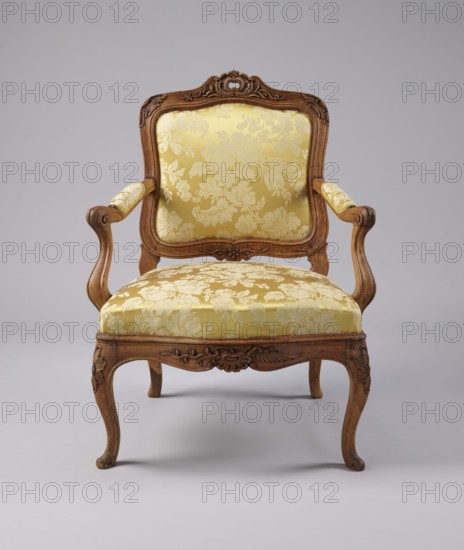 Armchair, rococo style, carved and upholstered seat, backrest and armrests, on four cabriole legs ending in turned-out slipper feet that arch into a wavy-cut, slightly bulging frame. Seat cushion recessed into the frame rebate on straps. S-shaped armrests and supports. Backrest disc-shaped, nearly rectangular, tilted outward with a clearance. Profiled frame elements enriched with carving: rosettes on the knees of the legs; on the front frame and horizontal backrest frames a rocaille motif framed by a plant twig and a bird's wing. Tenon joints reinforced with dowels; re-painted with gold paint. 3rd quarter of the 18th century, Italy, height 92 cm, width 70 cm, depth 64 cm., An ornate wooden armchair with carved frame and curved legs upholstered in a patterned fabric, showing a floral damask motif; the wood has a warm brown tone with carved decorative details on the crest rail, apron, arms, and knees, and the upholstery is a yellow-gold color with lighter yellow floral patterns; the chai...