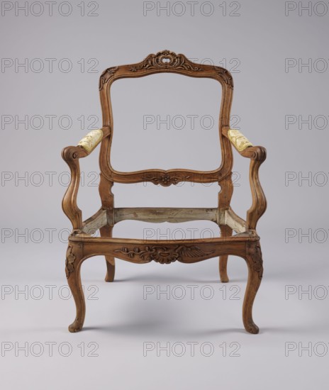 Armchair, rococo style, carved and upholstered seat, backrest and armrests, on four cabriole legs ending in turned-out slipper feet, arching into a wavy-cut, slightly bulging frame. Seat cushion recessed into the frame rebate on straps. S-shaped armrests and supports. Backrest disc-shaped, nearly rectangular, tilted outward with a clearance. Profiled frame elements enriched with carving rosettes on the knees of the legs; rocaille motif framed by a plant twig and a bird's wing on the front frame and horizontal backrest frames. Tenon joints reinforced with dowels; re-painted with gold paint. 3rd quarter of the 18th century, Italy, height 92 cm, width 70 cm, depth 64 cm., A wooden armchair frame with carved decorative details and curved legs sits against a light gray background; the wood shows warm brown tones with visible grain and carved floral motifs on the crest, apron, and knees, and the armrests feature a pale yellow patterned fabric on the top surfaces; the chair lacks seat and bac...