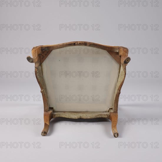 Armchair, rococo style, carved and upholstered seat, backrest and armrests, on four cabriole legs ending in turned-out slipper feet, arching into a wavy-cut slightly bulging frame. Seat cushion recessed into the frame rebate on straps. Armrests and supports are S-shaped. Backrest disc-shaped, nearly rectangular, tilted outward with a clearance. Profiled frame elements enriched with carving rosettes on the knees of the legs; on the front frame and horizontal backrest frames a rocaille motif framed by a plant twig and a bird's wing. Tenon joints reinforced with dowels; re-painted with gold paint. Ornament: shells, plant decoration. Rococo armchair, 3rd quarter of the 18th century, probably Italy. Seating piece, height 92 cm, width 70 cm, depth 64 cm., The image shows the back of an upholstered chair with a wooden frame, viewed against a pale gray background. The chair features a light beige fabric backing stretched across the frame, and the exposed wood has a worn, warm brown finish with...