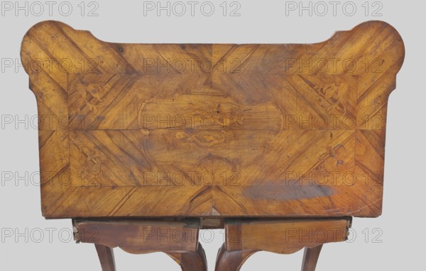 Game table, Rectangular with an extendable top square when extended with rounded, protruding corners, supported by four cabriole legs ending in hoof-shaped feet. Foot stained walnut; top veneered in walnut with inlay. Rear frame two-part: outer composed of two rotating frames on wooden hinges with legs attached on the inner side supporting the foldable top plate. Outer side of top framed by a crosswise frame with an asymmetrical, oval-shaped field, enclosed by channel bars and a ribbon, filled with floral and rocaille ornamentation. Inner side of tabletop has a wide walnut-veneered frame; center lined with green cloth. Game furniture, table; 1745–1770; Central Europe, possibly Saxony. Dimensions: height 74.6 cm, width 95 cm, depth 49 cm., A wooden tabletop seen from above with a warm brown and golden finish, showing wood grain patterns and a central decorative inlay with curved shapes and floral-like motifs; the tabletop has rounded corners on one side and straight edges on the other, ...
