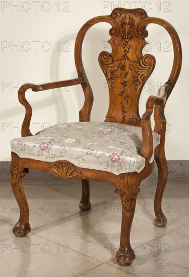 Armchair, animal paws, shells ornament, plant decoration, acanthus ornament, Neo-Baroque style, carved armchair with a padded seat, supported by four cabriole legs ending in a claw clasped around a ball. The knees of the legs and the front frame are decorated with carved shells. High, openwork backrest with a central board in the form of a vase, adorned with a carved plant ornament. Curved armrests repeating the outline of the seat, recessed, turning into S-shaped supports. Late 19th century, Central Europe. Piece of furniture, seating piece; height 105 cm, width 68.1 cm, depth 62.5 cm., A wooden armchair with carved detailing and a curving backrest, showing floral and scroll carving in the wood, featuring a patterned upholstered seat with a floral fabric in pale colors; visible colors include brown wood tones, cream, beige, pink, green, blue, and hints of yellow, set against a light beige wall and a grayish tiled floor.