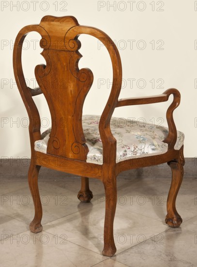 Armchair, animal paws, shells ornament, plant decoration, acanthus ornament, Neo-Baroque style. Carved armchair with a padded seat, supported by four cabriole legs ending in a claw clasped around a ball. The knees of the legs and the front frame are decorated with carved shells. High, openwork backrest with a central board in the form of a vase, adorned with a carved plant ornament. Curved armrests repeating the outline of the seat, recessed, turning into S-shaped supports. Late 19th century, Central Europe. Seating piece, height 105 cm, width 68.1 cm, depth 62.5 cm., A wooden armchair with a warm brown finish showing carved, curving lines and scroll details on the backrest and arms, a patterned upholstered seat in pale cream with floral motifs in muted colors including pink, blue, green, and beige, elegantly shaped cabriole legs ending in rounded feet, a polished stone or tiled floor in light gray and beige tones, and a plain off-white wall background.