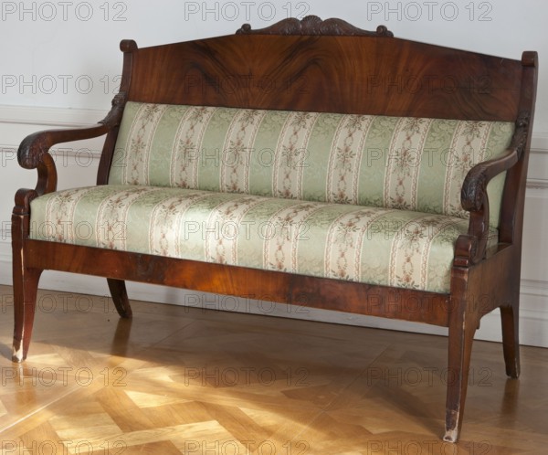 Sofa, Mahogany sofa with a padded seat and removable backrest cushions, on four sabre legs. Straight frame, wider at the sides. Backrest framed by angular S-shaped side frames, with a wide arched upper board topped with carved acanthus leaves. Rails finished with volutes on U-shaped supports; decorated with rosettes and palmettes. Biedermeier style, 1825–1840, possibly Vilnius. Seating furniture, height 104.7 cm, width 141.3 cm, depth 63 cm., A wooden settee with a polished dark brown backrest and carved decorative crest, curved carved armrests, and tapered legs, upholstered in a patterned fabric featuring vertical stripes and floral motifs in shades of pale green, cream, and muted pink, set against a light-colored wall and resting on a parquet floor with warm honey and golden brown tones.