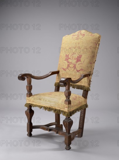 Armchair, neo-Baroque, padded seat and backrest upholstered with large-patterned silk fabric; supported by four legs fastened with an H stretcher — front legs baluster-shaped with a doll, rear legs angular and splayed outward. Armrest supports are angular, profiled balusters; S-shaped armrests ending in a front volute. High backrest flared upward with a wavy crest, straight in line with the sitter's back. Upholstery fabric symmetrical with stylized fleshy plant forms flowers, leaves, palmettes in cream on a red background; background has a satin warp weave, pattern formed by warp-faced threads. Frame at bottom and lower backrest trimmed with yellow silk fringe on a wide checkered ribbon; backrest sides and top trimmed with silver geometric braid. Backrest finished with yellow cotton grosgrain. Neo-Baroque style; 2nd half of the 19th century; Italy, / Austria ; seating piece armchair. Dimensions: height 121.5 cm, width 68 cm, depth 74 cm., An upholstered wooden armchair with a high back...