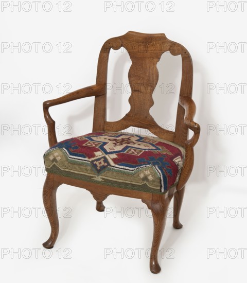 Children's armchair, origin unknown, rococo style, geometric ornament, cabriole legs, openwork backrests, for children. A children's armchair in the style of English Rococo furniture, with a trapezoidal padded seat and openwork backrest on cabriole legs ending in slipper feet. Narrow frame with bulging sides and a small front apron. S-shaped armrests with supports composed of two C-shaped beams. Side backrest slats are angular, topped with volutes framing the slightly rolled upper slat. Central backrest features a vase-shaped board mounted on a lower curtain frame. Seat on straps with later upholstery embroidered with crosses on canvas in a geometric pattern referencing oriental carpets; central is an isosceles cross in red, green, navy blue, and white. 3rd quarter of the 18th century; upholstery ca. 1870. Seating piece furniture. Height 71 cm, width 56.3 cm, depth 39.5 cm., An ornate wooden armchair with a carved backrest and curved arm supports, finished in a warm brown wood grain, f...
