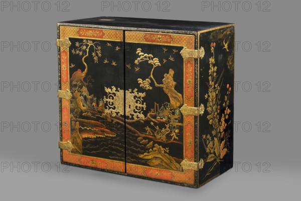 Cabinet, origin unknown, chinoiserie decoration with flowers, birds, trees, rocks, genre scenes against a landscape background, buildings, Chinese figures, bridges, geometric ornament, plant decoration; circa 1725, possibly Dresden; furniture storage cabinet; height 76 cm, width 85.5 cm, depth 41.5 cm., A rectangular wooden cabinet with a black lacquered surface decorated with gold-painted landscape scenes and floral motifs, featuring a central pair of doors with ornate gold metal hinges and an elaborate gold metal escutcheon; the doors and outer edges are framed with a decorative red border highlighted with gold patterns; the front shows stylized trees, rocks, birds, and architectural elements rendered in gold with touches of red and brown, while the side panel displays a vertical arrangement of flowers and foliage in gold with red accents; the top is plain black and the piece rests on a neutral gray background.