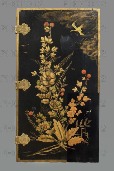 Cabinet, creator unknown, origin unknown, chinoiserie decoration with flowers, birds, trees, rocks, genre scenes against a landscape background including buildings and bridges, geometric ornament and plant decoration; ca. 1725; furniture storage cabinet; height 76 cm, width 85.5 cm, depth 41.5 cm., A tall rectangular black panel with a gold border features an intricate floral arrangement painted in gold with touches of orange, green, and dark blue, showing stylized blossoms, leaves, and stems rising from a base of elongated roots; a few round orange-red berries and small buds appear among the foliage, and several feathery fronds and fern-like leaves extend outward; near the top right a gold bird is depicted in flight against a clouded gold area, and a few small gold birds are scattered in the sky; ornate gold metal hinges are attached along the left edge, and the panel sits against a gray background.