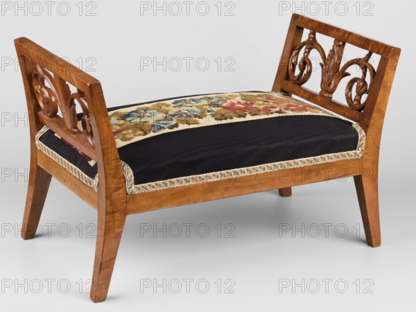 Footstool, Biedermeier style, decorated with flowers, plant motifs and acanthus tendril, circa 1820–1840, possibly Silesia place of origin, piece of furniture; H. 29 cm, W. 46.3 cm, D. 24.8 cm, A wooden bench with carved arm supports featuring scroll and leaf motifs, a padded seat with a patterned floral textile panel at the center and dark fabric on either side, decorative braided trim along the seat edge, and tapered wooden legs; visible colors include brown, black, beige, blue, red, green, and gold.