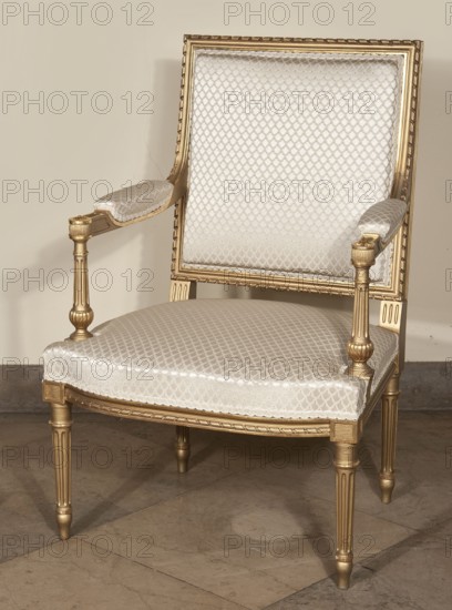 Armchair. Z. Szczerbinski i S-ka. Joint-Stock Company of the Warsaw Stylish Furniture Factory 1902–1939,  furniture factory author, neoclassicism style, balusters, Classicist-type armchair, carved, gilded, upholstered seat, backrest, armrests. Straight, turned, tapered, fluted legs with recessed profiles at the top separating the head, terminating in cubes. Trapezoidal seat, bulging at the front; rectangular backrest, straight in line with the back, with a clearance, tilted outward. Handrail supports in the form of turned balusters. Profiled frame elements; backrest and frame decorated with a spiral ribbon on the outside. Seat upholstery on springs. Early 20th century, Warsaw. Seating furniture, height 95.5 cm, width 63.5 cm, depth 60.5 cm., A decorative armchair with a wooden frame finished in gold and upholstered in a pale fabric with a diamond-like textured pattern, showing a rectangular backrest, cushioned seat, and padded armrests; the chair has turned and fluted legs and carved d...