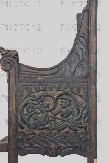 Armchair, wickerwork ornament, battle scenes, knights, plant ornament, dogs, ca. 1500 or 1st half of the 16th century, Sweden, Norway, piece of furniture, seating piece, height 112 cm, width 75 cm, depth 58 cm, The image shows a carved wooden panel with intricate raised reliefs and decorative molding, featuring a central human-like figure with flowing hair and a headband, seated or reclining and interacting with a stylized animal or serpent-like creature through a curved, ribbon-like form; surrounding the scene are swirling leaf and wave motifs, braided patterns, and scalloped edges. The wood displays a worn, aged surface with visible chips and scratches, and the predominant colors visible are dark brown and deep green with hints of muted reddish-brown where the finish is worn.