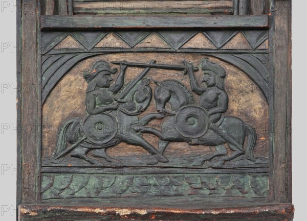 Armchair, wickerwork ornament, battle scenes, knights, plant ornament, dogs, ca. 1500 or 1st half of the 16th century, Sweden, Norway, piece of furniture, seating piece, height 112 cm, width 75 cm, depth 58 cm, A carved wooden relief panel shows a symmetrical scene of two mounted figures facing each other on horseback, each holding a long spear raised toward the other and carrying round shields at their sides; the riders wear distinctive helmets and have visible facial features and limbs, and the horses are rendered with bridles, manes, and raised legs suggesting motion. The carving is set within a rectangular frame with a curved arch above the scene and geometric triangular motifs in a band above; a carved leafy pattern runs along the bottom border. The surface shows layered paint and worn patina with visible colors including green, dark gray, brown, muted gold, and small areas of reddish-orange where the underlying material shows through.