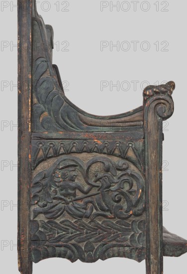 Armchair, wickerwork ornament, battle scenes, knights, plant ornament, dogs, ca. 1500 or 1st half of the 16th century, Sweden, Norway, piece of furniture, seating piece, height 112 cm, width 75 cm, depth 58 cm, A carved wooden panel showing a decorative side with a scroll-like armrest and ornate relief work featuring a human figure with wings holding a staff or spear and facing a stylized animal head entwined with curling vine-like forms, surrounded by layered wave and leaf motifs; the wood shows worn paint and surface wear with visible areas of green, dark brown, black, and touches of faded orange.