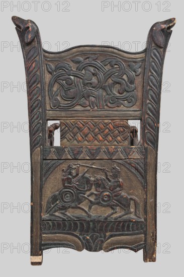 Armchair, wickerwork ornament, battle scenes, knights, plant ornament, dogs, ca. 1500 or 1st half of the 16th century, Sweden, Norway, piece of furniture, seating piece, height 112 cm, width 75 cm, depth 58 cm, A carved wooden chair back with a dark brown and muted black patina showing wear and touches of reddish-brown underlayer, featuring a top crest with two upward-facing stylized animal heads on each side, a central panel with intertwined vine and leaf scrollwork, a middle band with a woven lattice motif, and a lower panel carved in relief depicting a dynamic scene of two mounted figures engaged in combat with spears and round shields; the side stiles have twisted rope-like carved fluting, the lower apron has curved carved forms and a repeating scalloped motif, and the surface shows areas of lighter wood exposed where the finish is worn.