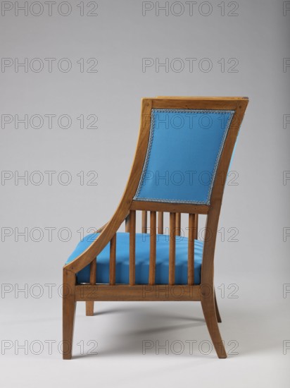 Armchair Berzera with a slender backrest and sides equal in height, arching upwards and forming ears, with angular frame elements. Front legs converge, rear legs arch outwards. Narrow, straight frame; trapezoidal seat in the form of a removable cushion on springs. The upper part of the sides and the backrest, which is slightly wider in the upper part, are upholstered; the lower part is filled with vertical slats. The frame elements are additionally connected with single pegs. Ca. 1820. Possibly Warsaw, Vienna, or Lesser Poland. Piece of furniture seating, height 86.5 cm, width 54.8 cm, depth 61.8 cm., A wooden chair shown in profile with a medium brown frame and a slatted lower back, a curved armrest and tapered legs, featuring blue upholstered panels on the seat and back with decorative trim, set against a neutral light gray background with soft shadow beneath.