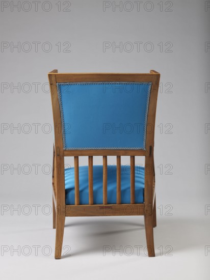 Armchair Berzera, pre-war collection. ARMCHAIR-BERZERA with a slender backrest and sides equal in height, arching upwards and forming ears, with angular frame elements. Front legs converge, rear legs arch outwards. Narrow, straight frame; trapezoidal seat in the form of a removable cushion on springs. The upper part of the sides and the backrest, which is slightly wider in the upper part, are upholstered, the lower part is filled with vertical slats. The frame elements are additionally connected with single pegs. Ca. 1820. Possibly Warsaw, Vienna, or Lesser Poland. Piece of furniture, seating piece. Height 86.5 cm, width 54.8 cm, depth 61.8 cm., A wooden chair viewed from the back with a rectangular upholstered back panel in bright blue trimmed with decorative edging, a matching bright blue cushioned seat visible through vertical wooden slats, and a light brown wooden frame and legs set against a neutral light gray background.