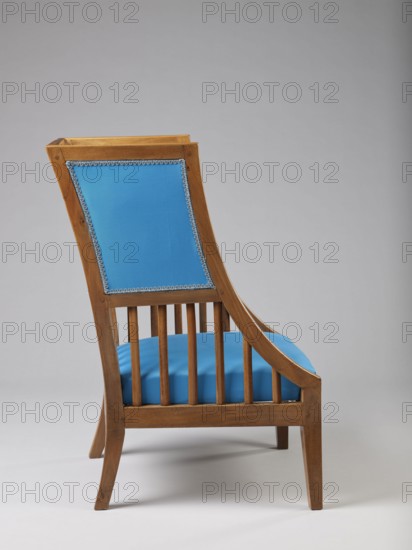 Armchair Berzera, armchair with a slender backrest and sides equal in height, arching upwards and forming ears, with angular frame elements. Front legs converge, rear legs arch outwards. Narrow, straight frame; trapezoidal seat in the form of a removable cushion on springs. The upper part of the sides and the backrest, which is slightly wider in the upper part, are upholstered; the lower part is filled with vertical slats. Frame elements are additionally connected with single pegs. Ca. 1820, possibly Warsaw, Vienna, or Lesser Poland. Piece of furniture seating, height 86.5 cm, width 54.8 cm, depth 61.8 cm., A wooden chair with a curved back and vertical slatted sides upholstered in bright blue fabric on the back panel and seat, the back panel edged with a decorative trim in a lighter color, the wood showing a warm brown tone with visible grain and smooth finish, set against a neutral light gray background and resting on a light gray floor.