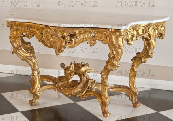 Console, origin unknown, flowers, rococo style, dragons, rocaille ornament, plant decoration, garlands. Console with a white marble top supported by a table-like, richly carved gilded base. The top and openwork frame have a wavy outline, bulge at the front, are rounded at the front corners, concave on the sides, and straight at the back. S-shaped legs fastened at the bottom with a cross-shaped lacing with wavy joints, with a winged dragon in the center. The structure is decorated with rocaille motifs, flowers, leaves, and channel sections. The frame is painted ochre on the inside. 1730–1750, Central Europe. Piece of furniture table. Dimensions: height 76 cm, width 143 cm, depth 72.8 cm., A decorative table with an ornate carved base finished in a metallic gold color and a smooth white top. The base features flowing curved legs and intricate scrollwork with floral and leaf motifs. The surface beneath the table shows a checkered floor with alternating dark and light squares. The backgrou...