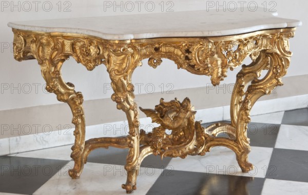 Console, origin unknown, flowers, rococo, dragons, rocaille ornament, plant decoration, garlands. Console with a white marble top supported by a richly carved, gilded base. Top and openwork frame have a wavy outline, bulging at the front, rounded front corners, concave sides, straight back. S-shaped legs joined at the bottom by a cross-shaped stretcher with wavy joints and a winged dragon at the center. Structure decorated with rocaille motifs, flowers, leaves, and channel sections. Frame painted ochre on the inside. 1730–1750, Central Europe. Piece of furniture, table; H 76 cm, W 143 cm, D 72.8 cm., A decorative table with a white marble top and an ornate gilded base featuring elaborate carved scrolls, floral motifs, and a sculpted winged creature, set against a pale wall and resting on a black and white checkered marble floor; visible colors include gold, white, black, beige, and hints of gray.