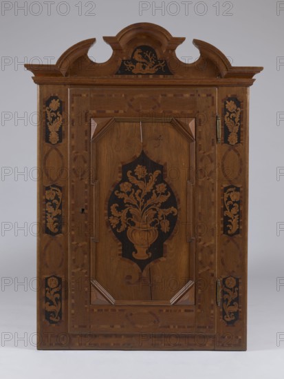 Built-in wardrobe front, baroque, rococo, ribbon-pincer ornament, flowers, vases with flowers, ca. 1762, Torun, fragment of furniture, height 138.5 cm, width 101 cm, depth 5.8 cm, A wooden cabinet door with a decorative shaped pediment at the top, featuring curved cutouts and an appliquéd curved crest, centered on a plain light gray background. The door front shows a central raised panel with angular corner moldings and an inlaid dark medallion containing a floral vase motif, surrounded by lighter wood grain. The frame around the central panel is detailed with patterned inlay borders and oval motifs on the stiles, and each side has vertical rectangular insets with floral marquetry. Visible hardware includes a metal hinge and a small escutcheon area. The overall color palette includes warm brown woods, darker brown or black inlaid areas, and subtle lighter brown highlights.