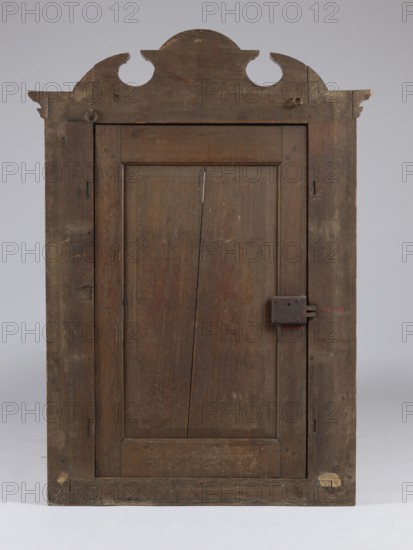Built-in wardrobe front, gift, baroque, rococo, flowers, ribbon-pincer ornament, vases with flowers, pre-war, ca. 1762, Torun, fragment of furniture, height 138.5 cm, width 101 cm, depth 5.8 cm, A tall wooden door with a decorative curved pediment at the top, showing worn and weathered brown wood tones throughout; the door has a recessed central panel with visible wood grain and a vertical split, a simple frame with darker brown stiles and rails, and an old rectangular metal latch on the right side; the surface displays scratches, scuffs, and patches of lighter and darker brown, with a few small areas of beige and pale worn spots near the bottom and top edges, all set against a neutral gray background.