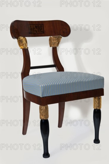 Chair, furnishings of representative buildings of the Republic of Poland 1918–1939, Administration of the Imperial Palaces in Warsaw 1815–1915, eagles, plant decoration, landscapes, Empire style. Mahogany chair with a padded seat removable cushion and an openwork backrest topped with an arched pelta-shaped board supported by a pair of eagles. The front legs are club-shaped, upper section carved with acanthus leaves; the rear legs are angular, curved outward in an arched manner. The seat and frame are straight at the front, arched at the back. The backrest rails are an extension of the rear legs, one-third of the height arched inward, fastened at the bottom with a turned black link. Early 19th century, Warsaw; seating furniture; height 83.1 cm, width 48 cm, depth 52 cm., A wooden chair with a dark brown curved backrest and dark brown frame, featuring carved ornamental blocks with gold-colored detailing where the back connects to the seat; the seat is upholstered in a striped fabric with...