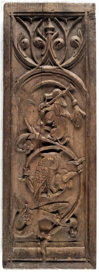 Furniture panel in bas-relief in the shape of an elongated standing rectangle, late 15th century, France. Fragment of furniture height 72.8 cm, width 25.2 cm, depth 3 cm. Decoration: plant twig with flowers bearing a dragon, an owl, and an animal playing bagpipes possibly a dog or monkey sitting on it; at the top tracery with fish-bladder motifs and a lily flower. Decoration enclosed in a rectangular frame with lines intersecting in the corners. Gothic style; drôlerie; animals, fantasy animals, plant ornament, musical instruments., A tall rectangular wooden panel carved with a vertical sequence of decorative motifs, showing a range of carved forms in brown wood tones. At the top a rounded arch framing stylized leaf and vine shapes with curved, pointed elements. Below that an arched frame contains a carved animal figure sitting upright holding an implement, with surrounding curved branches and floral motifs. Further down another circular frame encloses a carved bird with detailed feathe...