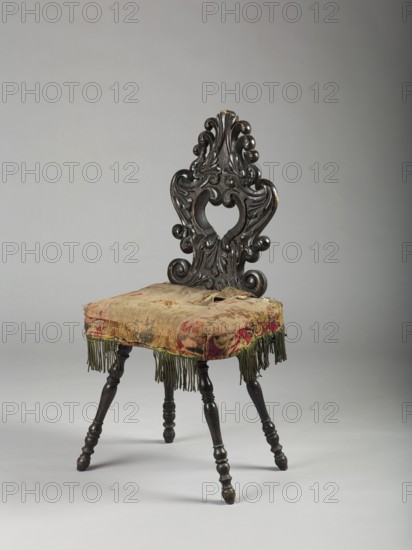 Stool with board seat, cushion attached, and openwork carved backrest; supported by four baluster legs splayed outwards. Backrest formed by a pair of connected acanthus volutes creating a heart-shaped opening. Seat trapezoidal with slightly concave sides and rounded corners. Cushion padded with horsehair and covered with sheared velvet, trimmed with fringe at the bottom. 3rd quarter of the 19th century, Southern Germany, Central Europe. Piece of furniture seating. Height 102 cm; width 49 cm; depth 42 cm., An ornate wooden chair with a dark brown carved backrest featuring scrolling, leaf-like motifs and a central open heart-shaped cutout, a worn upholstered seat covered in faded fabrics showing patches of red, green, and tan with frayed edges and hanging greenish fringe, and turned dark brown legs with rounded, bulbous shapes set against a neutral gray background.