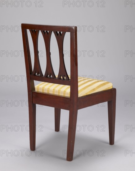 Chair, classicism style. Classicist chair with a trapezoidal padded seat insert cushion and rectangular openwork backrest filled with three lamellas, supported by four angular legs — front legs straight, rear legs arched outwards. Frame painted to look like mahogany painted imitation. Circa 1800, Poland . Piece of furniture; seating piece. Height 59 cm; width 36 cm; depth 36 cm., The image shows a wooden chair with a dark brown finish, viewed from a rear three-quarter angle against a light gray background; the chair has a rectangular backrest with decorative cutouts in the wood forming curved vertical shapes, straight tapered legs, and a seat cushion upholstered in a striped yellow and off-white fabric.