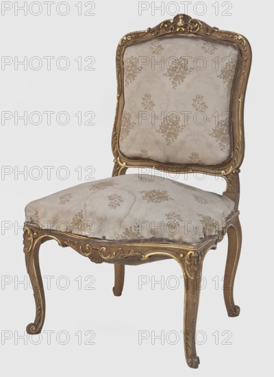 Chair, origin: Central Europe, ca. 1880. Neo-Rococo style with plant decoration and shell ornament. Rococo chair with wavy, carved, and gilded frame elements decorated with stylized acanthus leaves, palmette, and rocaille motif. Cabriole legs arching into the frame; legs terminate in volute feet. Trapezoidal seat upholstered on springs. Flat, disc-shaped backrest tilted outward with a clearance. Entire piece upholstered in beige silk grosgrain with floral bouquets. Seating piece; height 92 cm, width 56 cm, depth 55 cm., An upholstered chair with an ornate carved wooden frame finished in gold tones and brown, a patterned light beige fabric covering the backrest and seat with floral motifs in a slightly darker beige, curved legs with carved scroll and acanthus details in gold and brown, a padded seat with visible tufting and subtle wear, and a shaped backrest rimmed by the gilded carved frame.
