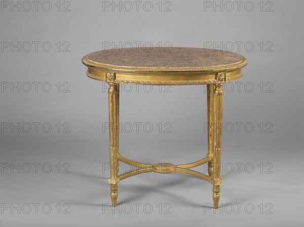 Table, classicism and Art Nouveau/Neoclassical styles, early 20th century, Central Europe, possibly Poland. Oval carved and gilded table with marble top, supported by four straight, angular tapering fluted legs joined with an X joint. Legs have inverted pear-shaped feet, arched at the top, ending in a cube decorated with a rosette; cubes are joined where the joints are set. Frame repeats the outline of the top, enclosed on both sides with a petal and at the bottom with a spiral ribbon motif. Stone tabletop slab enclosed in a wooden profiled frame decorated with a lesbian cymatium. Furniture table. Dimensions: height 77 cm, width 87.7 cm, depth 66 cm., An oval table with a brown mottled top set on a gilded frame; the frame and legs are gold colored with carved decorative details, fluted vertical legs connected by curved gold stretchers meeting at a round gold medallion, and the table stands on tapered gold feet against a neutral gray background.