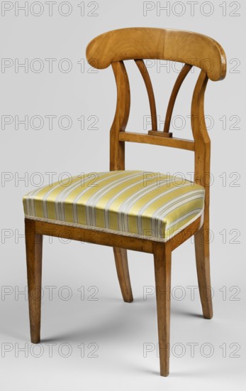 Chair, Biedermeier style, ca. 1825, Warsaw. Chair with an openwork backrest and upholstered seat on four angular, convergent legs: front legs straight, rear legs arch outward, flowing into the side stiles of the backrest. Seat rounded at the rear with a straight front frame. Backrest stiles arch inward at one-third of their height, joined at the bottom by an angular link and at the top by a wide board in the shape of an inverted pelta Amazonian shield. Upholstery attached to the frame. Seating furniture; height 91.2 cm, width 48.5 cm, depth 47.5 cm., A wooden chair with a curved top backrest and two curved vertical slats connecting to a horizontal support, a rectangular padded seat covered in striped fabric, and four straight tapered legs. The wood shows a warm honey-brown color with visible grain and some darker spots, the seat fabric features yellow and green stripes with pale accents, and the background is a neutral light gray.