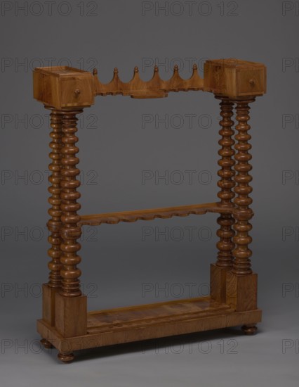 Pipe stand, origin unknown, smoking, pipes, acorns, rack for pipes and billiard cubes. Skeletal structure composed of two pairs of spool-turned columns on angular plinths, mounted on a narrow rectangular platform supporting cuboid drawer bodies, connected mid-height and at the top by horizontally arranged slats with semicircular cutouts on the front. Platform rests on four turned legs with a high, straight rim and glued-on wavy-cut slats for holding billiard cues or long pipes, and a single groove at the back probably originally from the installation of the gallery. Upper panel decorated with a gallery in the form of an arched crenellation reverse arcading topped with acorns. Bodies with drawers probably intended for balls are surrounded by a low slatted gallery. 1830–1840, Central Europe. Furniture, equipment; H 111.5 cm, W 89 cm, D 29 cm., A wooden freestanding rack with a warm brown finish, featuring a rectangular base with rounded feet, two turned vertical posts on each side with b...