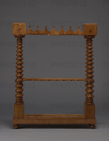Pipe stand, origin unknown, smoking, pipes, acorns, rack for pipes and billiard cubes. Skeletal structure composed of two pairs of spool-turned columns on angular plinths, mounted on a narrow rectangular platform, supporting cuboid drawer bodies connected mid-height and at the top by horizontally arranged slats with semicircular cutouts on the front. Platform rests on four turned legs with a high straight rim, with glued-on wavy-cut slats for holding billiard cues or long pipes, and a single groove at the back probably originally from installation. Upper panel decorated with a gallery in the form of an arched crenellation reverse arcading topped with acorns. Drawer bodies probably intended for balls are surrounded by a low slatted gallery. 1830–1840, Central Europe. Furniture/equipment. Height 111.5 cm, width 89 cm, depth 29 cm., The image shows a wooden freestanding rack with a warm brown finish against a neutral grey background; it has two tall turned vertical posts with a series of ...