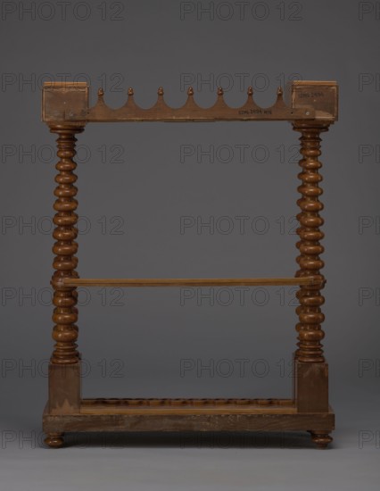 Pipe stand, origin unknown, smoking, pipes, acorns, rack for pipes and billiard cubes. Skeletal structure composed of two pairs of spool-turned columns on angular plinths, mounted on a narrow rectangular platform supporting cuboid drawer bodies, connected mid-height and at the top by horizontally arranged slats with semicircular cutouts on the front. Platform rests on four turned legs with a high straight rim, with glued-on wavy-cut slats for holding billiard cues or long pipes, and a single groove at the back. Upper panel decorated with a gallery in the form of an arched crenellation reverse arcading topped with acorns. Bodies with drawers probably intended for balls are surrounded by a low slatted gallery. 1830–1840, Central Europe. Furniture/equipment. Height 111.5 cm; width 89 cm; depth 29 cm., A wooden freestanding frame with a dark brown finish standing against a neutral gray background, featuring two turned vertical posts with a repeating bulbous profile connected by a horizonta...