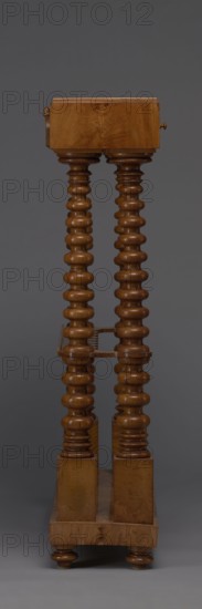 Pipe stand, origin unknown, smoking accessory featuring pipes and acorns; rack for pipes and billiard cubes. Skeletal structure of two pairs of spool-turned columns on angular plinths mounted on a narrow rectangular platform, supporting cuboid drawer bodies connected mid-height and at the top by horizontal slats with semicircular cutouts on the front. Platform rests on four turned legs with a high straight rim and glued-on wavy-cut slats for holding billiard cues or long pipes, with a single groove at the back. Upper panel decorated with a gallery in the form of an arched crenellation reverse arcading topped with acorns. Drawer bodies probably intended for balls are surrounded by a low slatted gallery. Dated 1830–1840, Central Europe. Furniture/equipment. Dimensions: height 111.5 cm, width 89 cm, depth 29 cm., A tall wooden piece of furniture with a warm brown finish stands against a dark gray background; it has a rectangular top block and a rectangular base connected by a pair of orna...