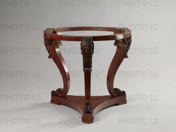 Table by Jakub Jan Simmler 1791–1872, carpenter. Round table supported by three S-shaped carved legs with acanthus volutes at both ends, mounted on a triangular platform with concave sides. Legs strongly flared at the top with half-cylinders at the rear edges, topped with volutes. Platform has three flat, block-shaped feet. Round, narrow frame glued from four layers of slats into a brick shape. Louis Philippe style, acanthus ornament, plant decoration, volutes. Dated 1840–1847. Furniture; table. Height 76.4 cm; diameter 93.5 cm., A wooden table base with a rich brown finish consisting of a circular top frame supported by a tripod of curved legs, each leg featuring carved decorative scrolls and leaf-like motifs near the top and bottom, and the legs meet a triangular stretcher base with carved accents at each corner; the surface shows glossy wood grain and polished highlights, and the background is a smooth light gray.