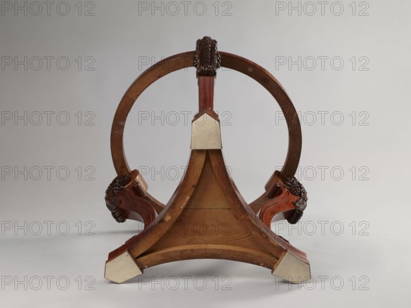 Table; maker: Simmler, Jakub Jan 1791–1872 carpenter. ROUND TABLE supported by three S-shaped carved legs, with acanthus volutes at both ends, mounted on a triangular platform with concave sides. The legs are strongly flared at the top, with half-cylinders at the rear edges, topped with volutes. The platform has three flat, block-shaped feet. Round, narrow frame, glued from four layers of slats into a so-called brick shape. Materials/ornament: acanthus ornament, plant decoration, volutes. Style: Louis-Philippe. Date: 1840–1847. Place of origin: Warsaw. Function/type: furniture; table. Dimensions: height 76.4 cm; diameter 93.5 cm., A wooden circular frame supported by a triangular wooden base is shown against a light gray background, with a group of ornate carved wooden elements at the junctions where the circular frame meets the base. The wood displays warm brown and reddish-brown tones with visible grain, and there are lighter beige protective coverings on the corners of the triangula...