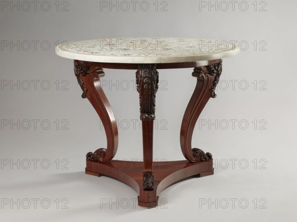 Table by Jakub Jan Simmler 1791–1872, carpenter. Round table supported by three S-shaped carved legs with acanthus volutes at both ends, mounted on a triangular platform with concave sides. Legs strongly flared at the top with half-cylinders at the rear edges, topped with volutes. Platform has three flat, block-shaped feet. Round, narrow frame glued from four layers of slats into a brick shape. Ornament: acanthus, plant decoration, volutes. Style: Louis Philippe. Function: furniture, table. Date: 1840–1847. Dimensions: height 76.4 cm; diameter 93.5 cm., A round table with a light-colored patterned top rests on a dark wooden base; the tabletop shows muted tones including pale, greenish, and reddish hues in a decorative design, and the edge appears creamy. The wooden base is a rich brown with a polished finish and features three curved legs joined to a triangular platform, each leg carved with ornate floral and scroll motifs in darker brown tones. The background is a smooth, neutral gray...