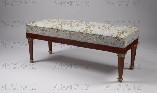 Banquette, Empire style, early 19th century, Warsaw, rectangular mahogany banquette, upholstered, supported by four turned, tapering legs with brass capitals and feet. Straight polished frame with cast, chiseled, gilded fittings. Spring-mounted upholstery. Dimensions: height 52 cm, width 122.5 cm, depth 48 cm., A rectangular padded bench with a patterned fabric top in pale blue and gold rests on a wooden frame in warm brown; the fabric shows ornate floral and medallion motifs in gold on the blue background. The wooden skirt is brown and the tapered legs are brown with gold-colored metal caps and collars. The bench sits on a smooth light gray surface against a neutral gray backdrop, and there are soft shadows beneath the bench. The visible colors include pale blue, gold, brown, light gray, and neutral gray.