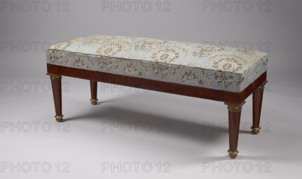 Banquette, Empire style, early 19th century. Rectangular mahogany banquette, upholstered, supported by four turned, tapering legs with brass capitals and feet. Straight polished frame with cast, chiseled, gilded fittings. Spring-mounted upholstery. Dimensions: height 52 cm, width 122.5 cm, depth 48 cm. Origin possibly Warsaw., A rectangular upholstered bench with a patterned light blue fabric top featuring ornate floral and medallion motifs in gold and beige sits on a polished wooden frame in a warm brown tone; the bench has a decorative trim along the edge of the upholstery and tapered wooden legs with brass or gold-colored accents at the tops and feet, all set against a neutral gray background and casting soft shadows on a pale gray floor.