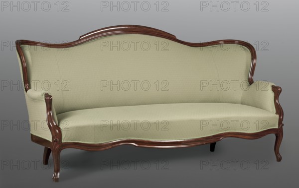 Sofa, Office of the Council of Ministers provenance, Ludwik Filip style, mahogany. SOFA, carved, upholstered seat, backrest, inner and outer sides with armrests, on four low, cabriole legs. The frame has a wavy line, emphasized by a half-cylinder. The backrest is rounded on the sides, slightly curved in the line of the back and tilted outwards. The armrest supports are S-shaped. The whole is upholstered in crimson plush. 1850–1870, Warsaw. Piece of furniture, seating piece. Height 111 cm, width 216 cm, depth 65 cm., A vintage-style sofa upholstered in a light green fabric with a subtle repeating pattern is shown against a neutral gray background, featuring a carved wooden frame in a rich brown finish that outlines the curved backrest, arms, and base; the seat is a single continuous cushion and the arms are rolled with the wood visible at the front, and the sofa stands on sculpted wooden legs.