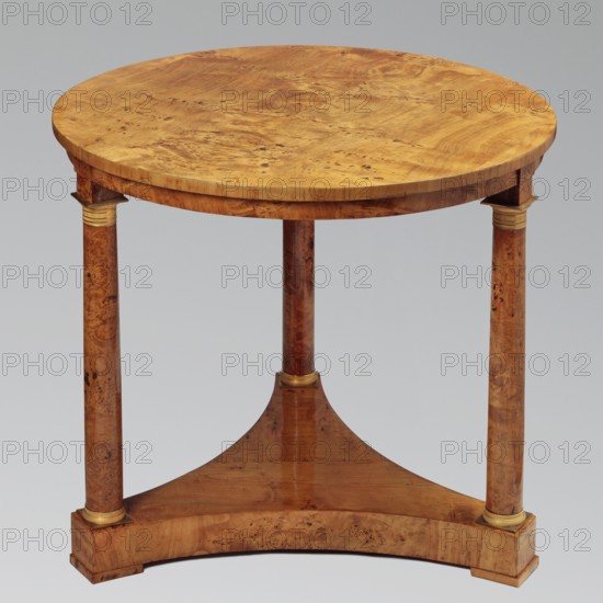 Round table on three legs in the form of slender columns with smooth shafts, mounted on a triangular platform with concave sides. Entire piece veneered with poplar burl. Cube feet at the corners of the platform. Bases and capitals of the columns are cast brass, covered with a twisted rope motif and stylized leaves. Biedermeier style, 1815–1825, Warsaw. Piece of furniture; height 75.8 cm, diameter 84 cm., A round wooden table with a warm golden-brown surface and visible wood grain sits on three cylindrical legs that have a slightly darker brown wood tone and natural wood patterning; a triangular wooden stretcher in a medium brown connects the legs and rests on a curved wooden base piece that matches the tabletop's golden-brown color, and there are small decorative bands of pale gold at the top and bottom of each leg.