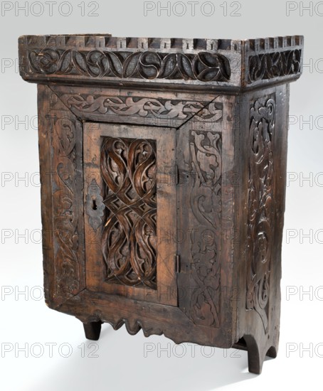 Hanging cabinet, ca. 1500, Germany Bavaria? Tyrol? — Gothic style. Wooden hanging cabinet with straight sides and front, topped by a prominent crenellated cornice; closed by a single door with a single narrow bottom shelf, carved in bas-relief. Lower-section sides extended with wavy ends. Door engraved with plant twigs at top and sides; door leaf and baseboard covered with sharply cut fish-bladder ornament and dry thistle ornament. Sides, front, and back composed of boards glued together and reinforced with dowels. Interior with one shelf. Steel fittings: push-lock without cup, braided hinges, and a club with ring mounted in the upper middle of the back. Furniture; storage cabinet. Height 65.3 cm, width 45 cm, depth 29.2 cm., A dark brown wooden cabinet with carved decorative panels stands against a light grey background, showing a rectangular top with carved border motifs, a front door with an openwork central panel featuring flowing interlaced carvings, carved side panels with simila...