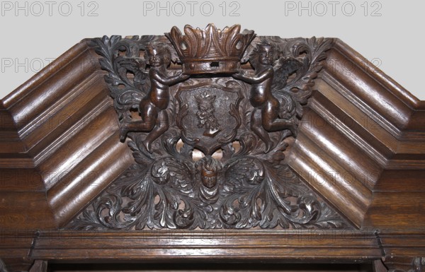 Wardrobe, putti, Baroque style, coats of arms, plant decoration, lion masks, personifications of the seasons: Spring, Summer, Autumn, Winter; dated 1710–1730, probably Gdansk, Royal Prussia; piece of furniture storage, height 200 cm, width 124 cm, depth 62 cm., A carved wooden panel featuring a central shield topped by a crown held by two winged cherubic figures, with ornate acanthus leaf scrollwork surrounding the shield and filling the lower area where a single cherubic head with wings appears; the carving and surrounding frame show a polished dark brown wood with highlights of lighter brown from reflected light, and a neutral pale gray background is visible above the panel.
