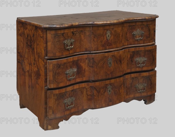 Chest of Drawers, Baroque, floral decoration. Chest of Drawers with straight sides and a wavy front, three drawers, supported by four console legs, veneered with patterned walnut. Drawer faces and top with frames. Brass openwork wavy-cut fittings. Circa 1750, Central Europe. Piece of furniture, storage unit. Height 85 cm, width 121 cm, depth 64 cm., A wooden chest of drawers with a curved front and carved base, showing a rich brown wood grain with darker brown and reddish-brown variations, ornate metal handles and keyhole escutcheons in a muted metallic tone, a smooth flat top surface, and visible wood joints and edge molding; the background is a pale gray.