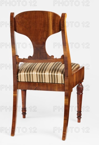 Chair, acanthus ornament, Biedermeier style, palmettes ornament. Mahogany chair with a padded seat removable cushion and a board backrest. Front legs straight, turned in the form of a baluster; rear legs sabre-shaped. Trapezoidal seat, slightly bulging at the front. Wide side frames embrace the seat cushion. The cushion rests on a pine frame, upholstered in green-striped fabric. The backrest consists of angular, S-shaped side stiles, a lower crossbar, and a rounded central board placed between them, narrowed at the bottom, straight in line with the back. The backrest stiles are widened in the lower section, ending at the top with a forward-curved volute. Polished frame enriched with carving — acanthus leaves on the upper part of the front legs, antemion and symmetrical S-shaped beams at the bottom of the backrest boards, scrollwork at the ends of the backrest crossbars. 1830–1840, Warsaw, Poland. Seating furniture; furniture. Height 92 cm, width 51.4 cm, depth 56.5 cm., A wooden chair ...