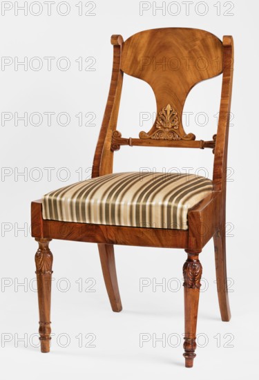 Chair, Biedermeier, mahogany chair with a removable padded seat cushion and board backrest. Front legs straight, turned in the form of a baluster; rear legs sabre-shaped. Trapezoidal seat, slightly bulging at the front. Wide side frames embrace the seat cushion, which rests on a pine frame upholstered in green-striped fabric. Backrest with angular S-shaped side stiles, a lower crossbar, and a rounded central board narrowed at the bottom and straight in line with the back. Backrest stiles widened in the lower section, ending at the top with a forward-curved volute. Polished frame enriched with carving—acanthus leaves on the upper part of the front legs, antemion and symmetrical S-shaped beams at the bottom of the backrest boards, and scrollwork at the ends of the backrest crossbars. Palmettes ornament. Dated 1830–1840, probably Warsaw, Poland. Seating furniture; height 92 cm, width 51.4 cm, depth 56 cm., A wooden chair with a curved backrest and carved decorative element on its lower ba...