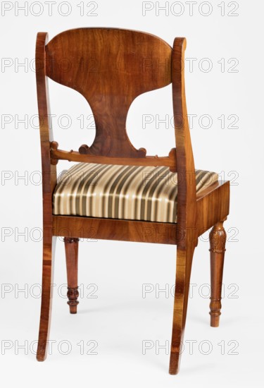 Chair, Biedermeier style, mahogany; padded seat with removable cushion and board backrest. Front legs straight, turned in baluster form; rear legs sabre-shaped. Trapezoidal seat, slightly bulging at the front. Wide side frames embrace the seat cushion, which rests on a pine frame upholstered in green-striped fabric. Backrest has angular S-shaped side stiles, a lower crossbar, and a rounded central board narrowed at the bottom and aligned with the back. Backrest stiles widen in the lower section and end with a forward-curved volute. Polished frame with carving: acanthus leaves on the upper front legs, anthemon antemion and symmetrical S-shaped beams at the base of the backrest boards, and scrollwork at the ends of the backrest crossbars. Dated 1830–1840, possibly Warsaw, Poland. Seating furniture; height 92 cm, width 51.4 cm, depth 56 cm., A wooden chair seen from the back with a curved top rail and a sculpted splat showing wood grain in warm brown tones, a horizontal stretcher connecti...