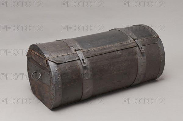 Trunk, A TUBE in the shape of a cylinder truncated at the base, covered with thick leather and upholstered with four steel straps, fastened with steel heads, with U-shaped handles on the sides of the body. The lid is closed with two hasps, attached to strap hinges. The connection between the body and the lid is protected from water by a leather apron. The side straps on the outside have semicircular cutouts, folded. The interior of the trunk is lined with gray canvas. The lid, when opened, is held on the sides with leather strips. 17th/18th century, Poland, piece of furniture, height 46 cm, width 104.5 cm, depth 51.5 cm. /creator; deposit provenance, pre-war., A cylindrical chest with a rounded top made of dark metal showing worn and rough textures sits on a neutral light gray surface; the chest has a few large metal bands encircling it and several rivets along seams, with two metal latch straps on top and a metal handle on one end; visible colors include dark gray, black, brownish rus...