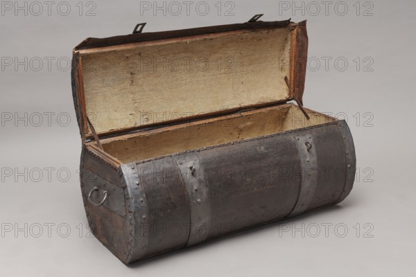 Trunk. A cylindrical-truncated tube covered with thick leather and reinforced with four steel straps fastened with steel heads, with U-shaped handles on the sides. Lid closed with two hasps attached to strap hinges; connection between body and lid protected from water by a leather apron. Side straps on the outside have semicircular folded cutouts. Interior lined with gray canvas; lid held open by leather strips. 17th/18th century, Poland, piece of furniture; H 46 cm, W 104.5 cm, D 51.5 cm., An open, curved-top trunk with a dark brown exterior reinforced by metal bands and metal rivets; the trunk has a worn, textured surface and metal handles on its sides. The interior shows a coarse, light beige fabric lining covering the lid and inside walls, with some small stains and fraying. Leather straps and metal fixtures are visible at the lid hinges and edges, and the trunk rests on a smooth, pale gray background. The visible colors include dark brown, beige, gray, and hints of rust-toned brow...